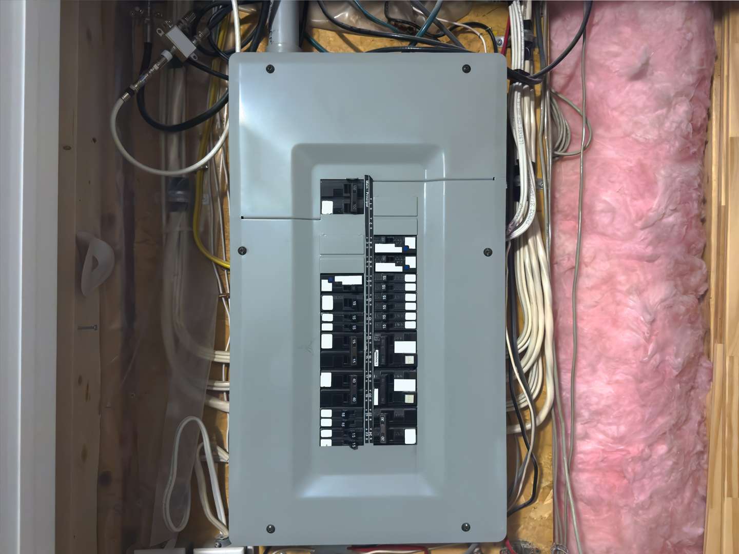 Residential electrical breaker panel with multiple circuit breakers, wires organized on both sides, and pink insulation visible on the right.